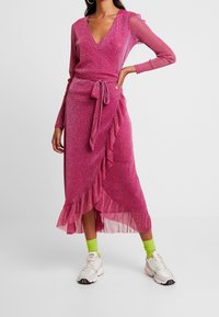 Vestido envolvente rosa brillante con mangas largas, dobladillo con volantes y detalle de cintura anudada, combinado con zapatillas blancas y calcetines verdes.