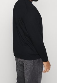 Svart stickad tröja med långa ärmar, rund halsringning och ribbad fåll. Bärs tillsammans med grå denimbyxor och visar en sidovy från midjan och uppåt.