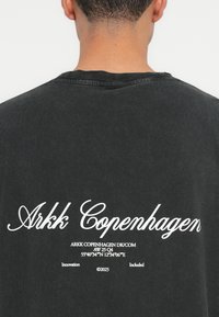 Crna pamučna majica s okruglim izrezom, s bijelim tipografskim natpisom na stražnjoj strani, koji prikazuje "Arkk Copenhagen" i dodatni tekst.