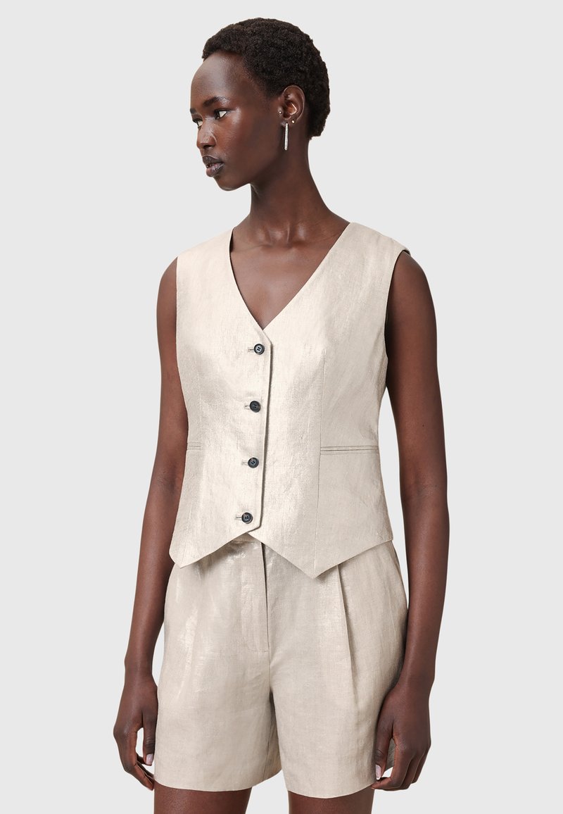 Beige linneväst med v-ringning, fem svarta knappar och en åtsittande design. Bärs över matchande plisserade shorts med slät textur och skräddarsydd form.