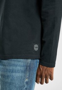 Zwarte longsleeve shirt met geribbelde textuur, met een logo patch nabij de zoom, gecombineerd met blauwe spijkerbroeken.