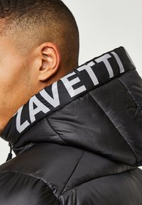 Schwarze Daunenjacke mit hohem Kragen und "ZAVETTI" in Silber aufgedruckt. Das Material ist glänzend und hat eine gesteppten Textur.