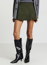 Minigonna in velluto a coste verde oliva con spacco laterale, abbinata a stivali neri in vernice aderenti fino al ginocchio con punte appuntite.
