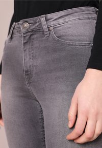 Primo piano di una persona che indossa jeans grigi attillati e una maglietta nera a maniche lunghe, con la mano destra poggiata al fianco.