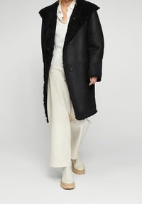 Abrigo de borrego negro con un amplio collar, dos botones y bolsillos laterales; combinado con una blusa blanca y pantalones de pierna ancha color crema.