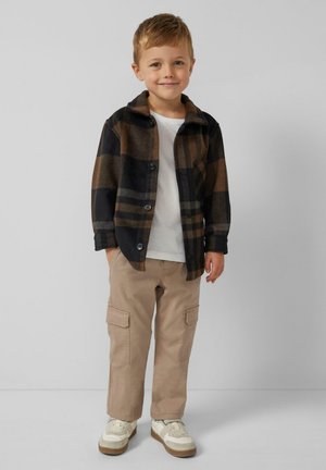 Dziecko ubrane w czarną i brązową koszulę w kratę z flaneli, białą koszulkę, beżowe spodnie bojówki i białe trampki z brązowymi akcentami.