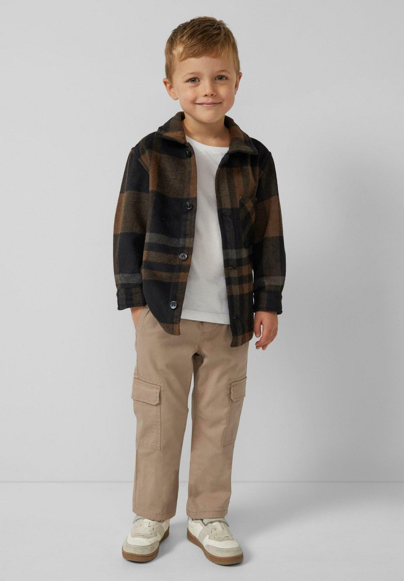 Dziecko ubrane w czarną i brązową koszulę w kratę z flaneli, białą koszulkę, beżowe spodnie bojówki i białe trampki z brązowymi akcentami.