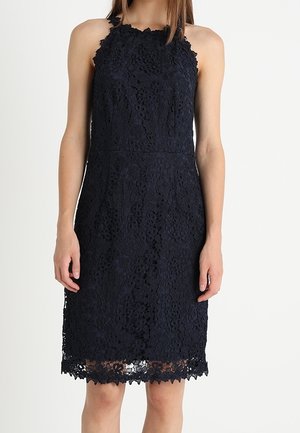 Mouwloze zwarte kanten jurk tot op de knie met bloemenpatroon, getailleerde taille en geschulpte zoom, gedragen door een staand persoon.