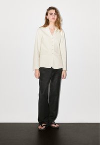 Frau steht vor einer schlichten weißen Wand, trägt einen cremefarbenen, geknöpften Blazer, lockere schwarze Hosen und schwarze Flip-Flops, mit lockigem Haar über den Schultern.