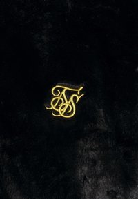Sort faux fur stof med en blød tekstur har et fremtrædende gult broderet monogram i et udsøgt skrift design.