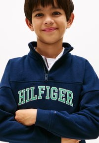 Μπλε σκούρο φούτερ με φερμουάρ και την επιγραφή "HILFIGER" σε πράσινα γράμματα. Διαθέτει ψηλό γιακά και ριμπ μανσέτες. Μαλακή υφή υφάσματος.