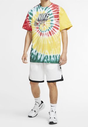 Osoba nosząca wielokolorową koszulkę Nike tie-dye z grafiką, białe spodenki do koszykówki, białe skarpetki Nike i białe trampki, stojąca.