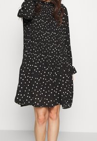 Svart klänning med långa ärmar, löst snitt och rynkad halsringning, prydd med vita prickar över hela tyget.