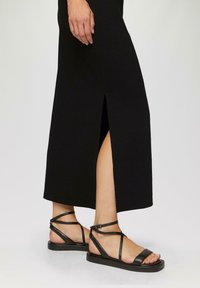 Robe maxi noire avec une fente sur le côté, tissu texturé. Sandales en cuir noir chunky avec des brides croisées et une fermeture à boucle.