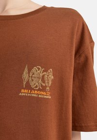 Braunfarbenes Baumwoll-T-Shirt mit kurzen Ärmeln. Mit einem hellgrünen Grafikdesign von einer Muschel, einem Krebs und einem Fisch, sowie der Aufschrift "BILLABONG ADVENTURE DIVISION" in Orange.