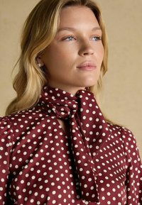 Maroon blouse with white polka dots, featuring a tied scarf neck detail, long sleeves, and a smooth fabric texture.