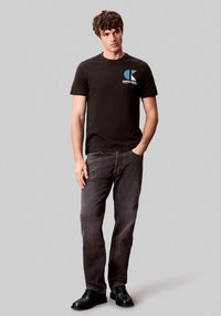 Camiseta negra de manga corta con un logo de Calvin Klein azul y blanco, combinada con jeans de pierna recta gris oscuro y zapatos negros.