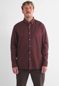 Chemise à manches longues bordeaux avec un col boutonné et des boutons blancs. Présente un petit logo bleu marine sur la poitrine, fabriqué en tissu lisse.