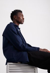 Camicia button-up blu navy in una vestibilità rilassata, realizzata in cotone. Presenta un colletto, maniche lunghe e un'etichetta con logo sull'orlo. Indossata con pantaloni neri.