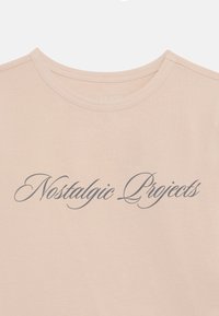 Heled roosa T-särk, mis on valmistatud puuvillast, tumeda halli käekirjaga "Nostalgic Projects" trükiga rinna peal. Ümmargune kaeluse disain.