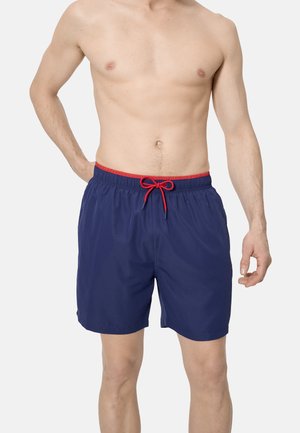 Hombre con pantalones cortos de baño azul marino con cordón rojo, de pie contra un fondo liso, mostrando la sección media y las piernas superiores.