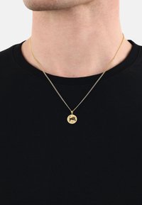 Guldkedja med en cirkulär berlock med en tjurdesign, hängande från en fin kedja, bar över en svart t-shirt.