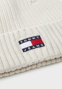 Knit beige beanie with ribbed texture. Features a rectangular logo patch in navy, red, and white with "TOMMY JEANS" text.