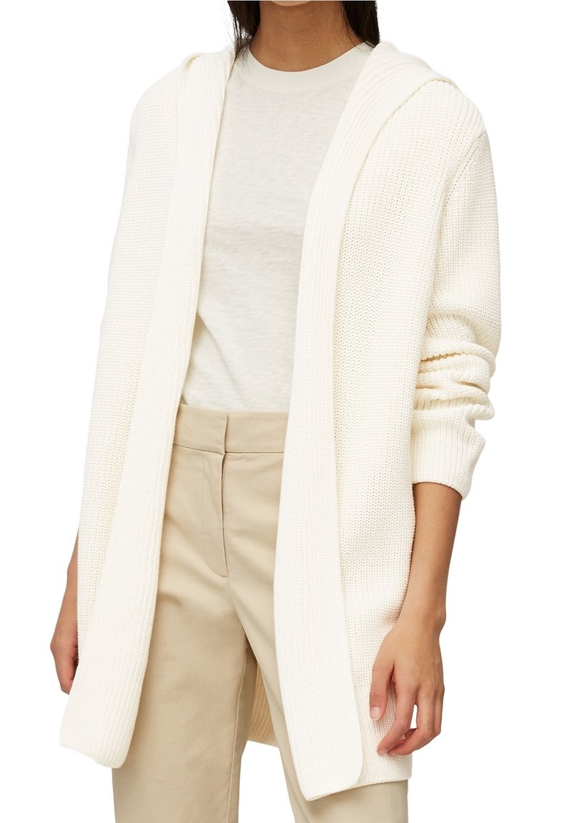 Žena v krémovém pleteném kardiganu s otevřeným předním dílem a kapucí, s dlouhými rukávy a béžovými kalhotami s vysokým pasem, stojí s uvolněnými pažemi.