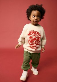 Crème sweatshirt met rode "Little Monster Racing Champions" grafiek, gecombineerd met olijfgroene cargobroek en witte sneakers. Bruine achtergrond.