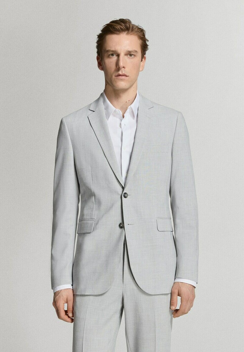 Man draagt een lichtgrijs colbert met twee knopen en bijpassende broek, met een wit overhemd, staand tegen een effen lichte achtergrond.