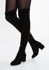 Piernas vistiendo medias opacas negras y botas de tacón de ante negras por encima de la rodilla, cruzadas contra un fondo blanco.