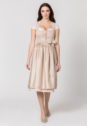 Frau trägt ein beiges, knielanges traditionelles Kleid mit Spitzenbluse und einer mit einer seitlichen Schleife gebundenen Satinschürze, stehend vor einfachem Hintergrund.