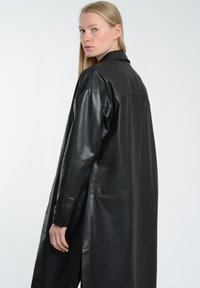 Sort læderfrakke med lange ærmer, skjortekrave og sideslids. Glat tekstur og en afslappet pasform, der fremhæver minimalistisk design.