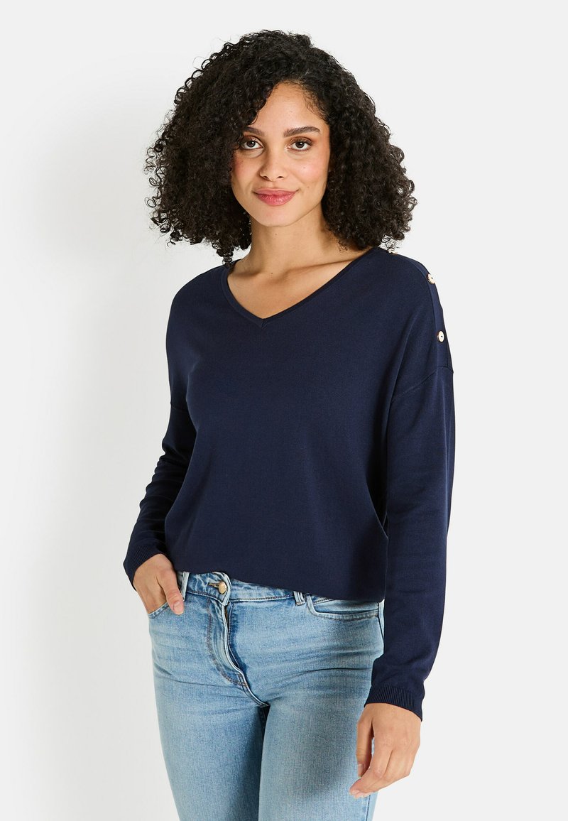 Mujer con cabello rizado vistiendo un suéter navy de cuello en V con botones en el hombro y jeans azul claro, de pie con una mano en el bolsillo.