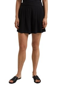 Mustat laskostetut shortsit, joissa sileä kangas, istuva vyötärönauha ja rento istuvuus. Yhdistetty mustiin litteisiin liuku-sandaaleihin.