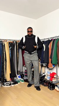 Schwarze Collegejacke mit weißen Akzenten, graue gestreifte Hose und schwarze Schuhe. Der Hintergrund zeigt verschiedene Kleidung und Schuhe auf Ständern.