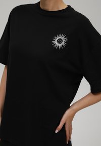 Crna pamučna majica sa bijelim grafičkim motivom sunca i mjeseca na prsima, okrugli izrez, kratki rukavi i opušteni kroj.