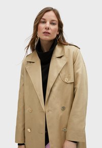 Beige double-breasted coat with subtle embossed patterns, large lapels, and two button closure. Worn over a black turtleneck.