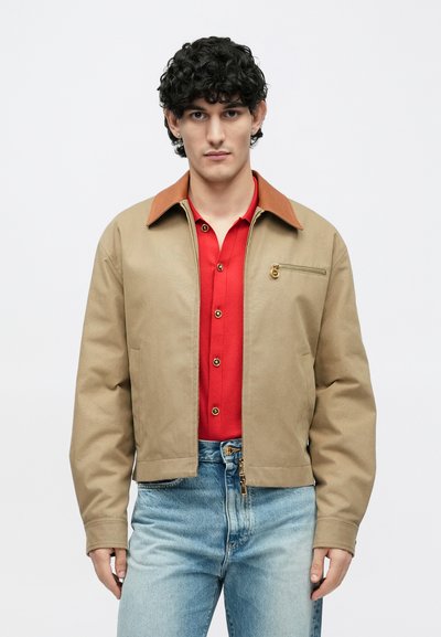 Vyras su garbanotais tamsiais plaukais, vilkintis smėlio spalvos striukę su rudais atvartais, raudoną marškinių su sagutėmis, šviesiai mėlynas džinsines kelnes ir auksinius auskarus, fotografuotas prieš paprastą foną.