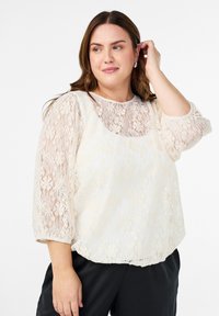 Sieviete, kas pielāgo matus, ģērbta kremines krāsas mežģīņu blūzē ar trīs ceturtdaļu piedurknēm un melnā biksēs, stāv uz baltas fona.