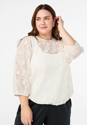 Kobieta poprawiająca włosy, ubrana w kremową koronkową bluzkę z rękawami trzyczwartowymi i czarne spodnie, stojąca na białym tle.