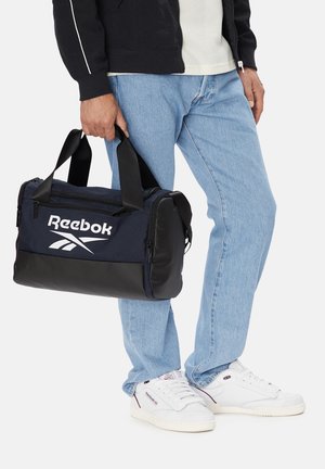 Osoba ubrana w jasnoniebieskie dżinsy i białe buty Reebok trzyma torbę sportową Reebok w kolorze granatowo-czarnym za uchwyty.