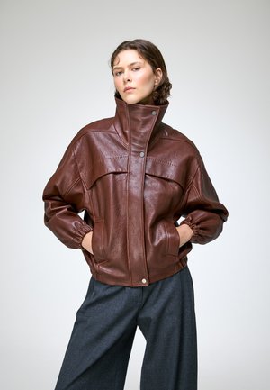 Jeune femme portant une veste en cuir marron à col montant et poignets élastiques, les mains dans les poches, et un pantalon ample gris foncé sur fond uni.