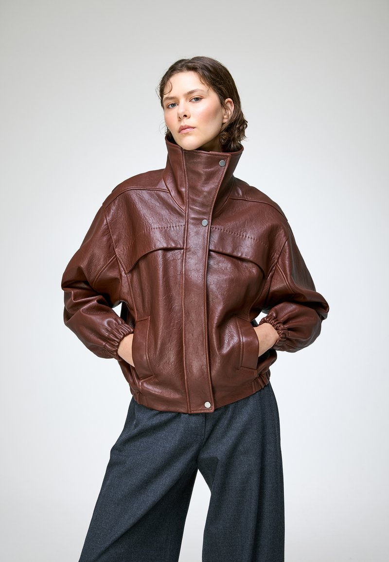 Jeune femme portant une veste en cuir marron à col montant et poignets élastiques, les mains dans les poches, et un pantalon ample gris foncé sur fond uni.