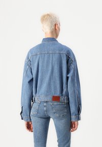 Giacca di jeans azzurro chiaro con finitura testurizzata, vestibilità casual, spalle scese e toppa in pelle sul retro. Dettagli con polsini abbottonati.
