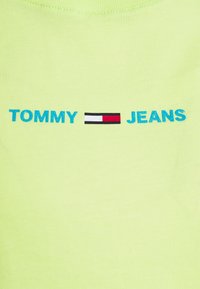 Neongrön t-shirt med ribbad textur. Har texten "TOMMY" i blått och "JEANS" i blått, åtskilda av en liten svart, röd och vit logoblock.