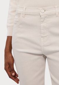 Jeans de um bege claro com uma textura suave, apresentando bolsos padrão, fecho com botão e detalhes de costura discretos ao longo das costuras.