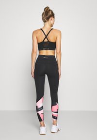Sort sports-bh med krydsede stropper; sorte leggings med pink og sorte geometriske mønstre; parret med hvide atletiksko.