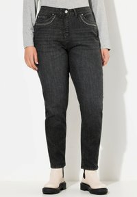 Jeans in denim nero con una forma aderente, cuciture decorative sui taschini anteriori e una leggera lucentezza, indossati con stivali alla caviglia bianco sporco.