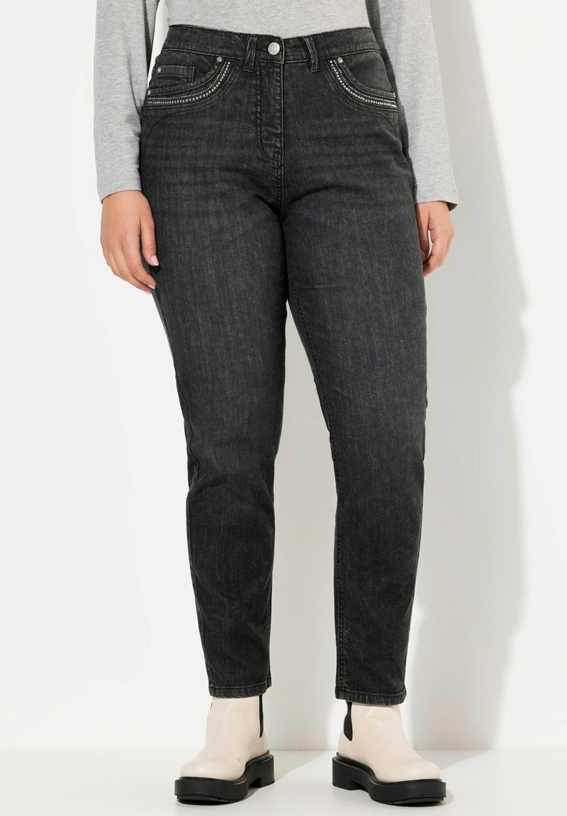 Jeans in denim nero con una forma aderente, cuciture decorative sui taschini anteriori e una leggera lucentezza, indossati con stivali alla caviglia bianco sporco.
